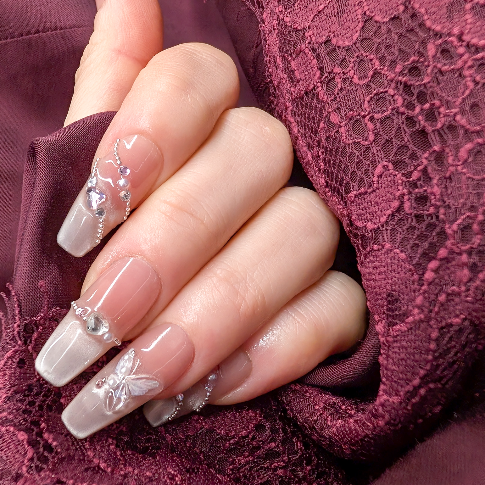 A close-up of a hand with long, glossy coffin-shaped nails decorated with clear rhinestones and a butterfly motif, resting against deep burgundy lace fabric.