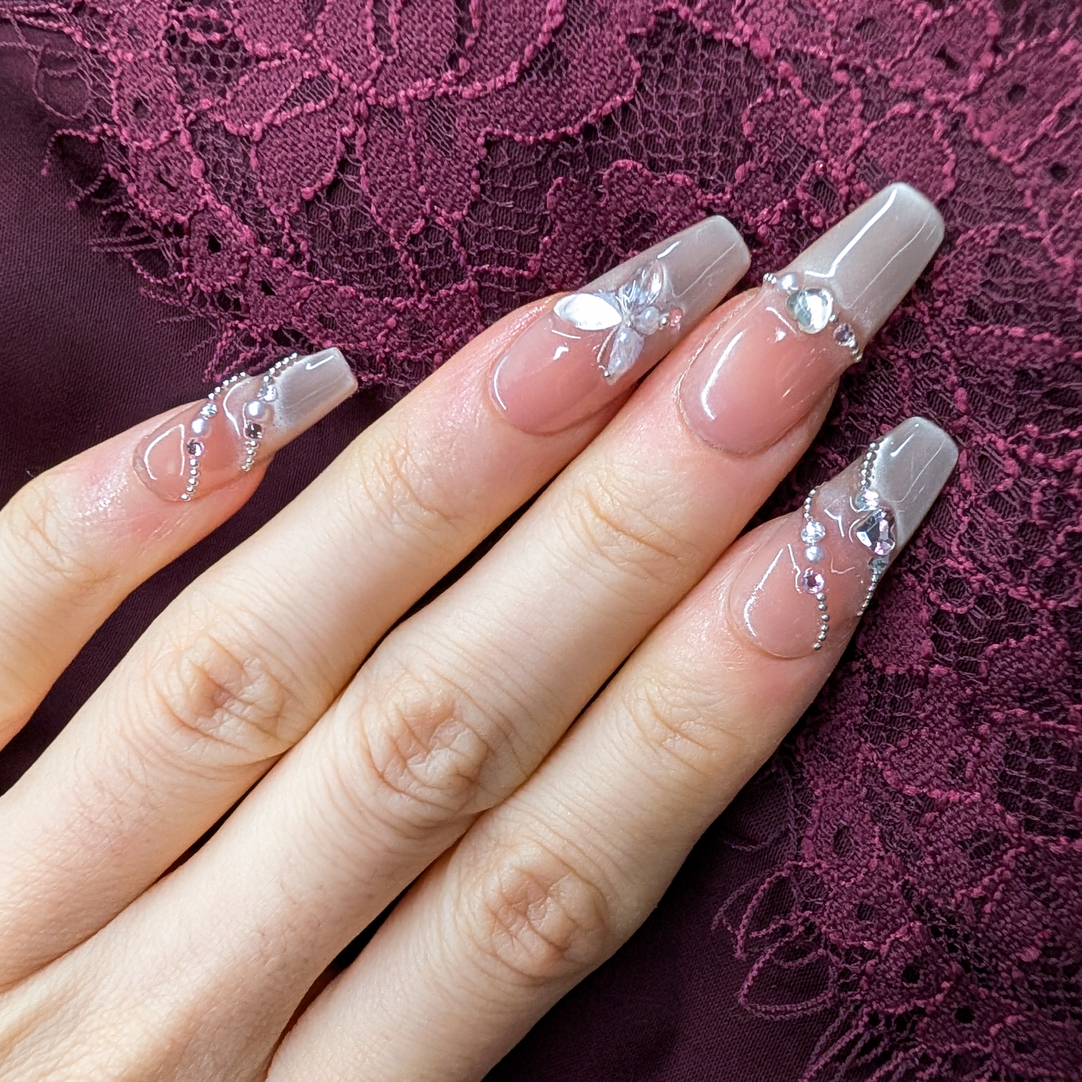 A close-up of a hand with long, coffin-shaped press-on nails featuring a glossy nude-pink base, clear glittery tips, and silver butterfly and rhinestone nail art, set against a dark purple lace fabric background.