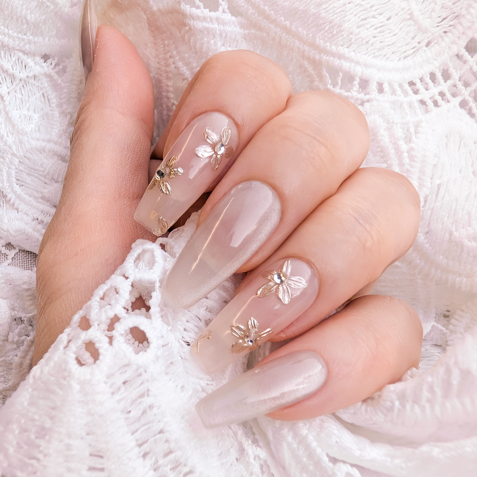 A close-up of a hand with long, glossy almond-shaped nails decorated with delicate gold floral and butterfly designs, resting on white lace fabric.