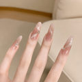 A close-up of a hand with long, almond-shaped nails featuring a sheer nude-pink gradient, white feather-like brushstroke details, and small clear rhinestone accents, set against a soft beige indoor background.