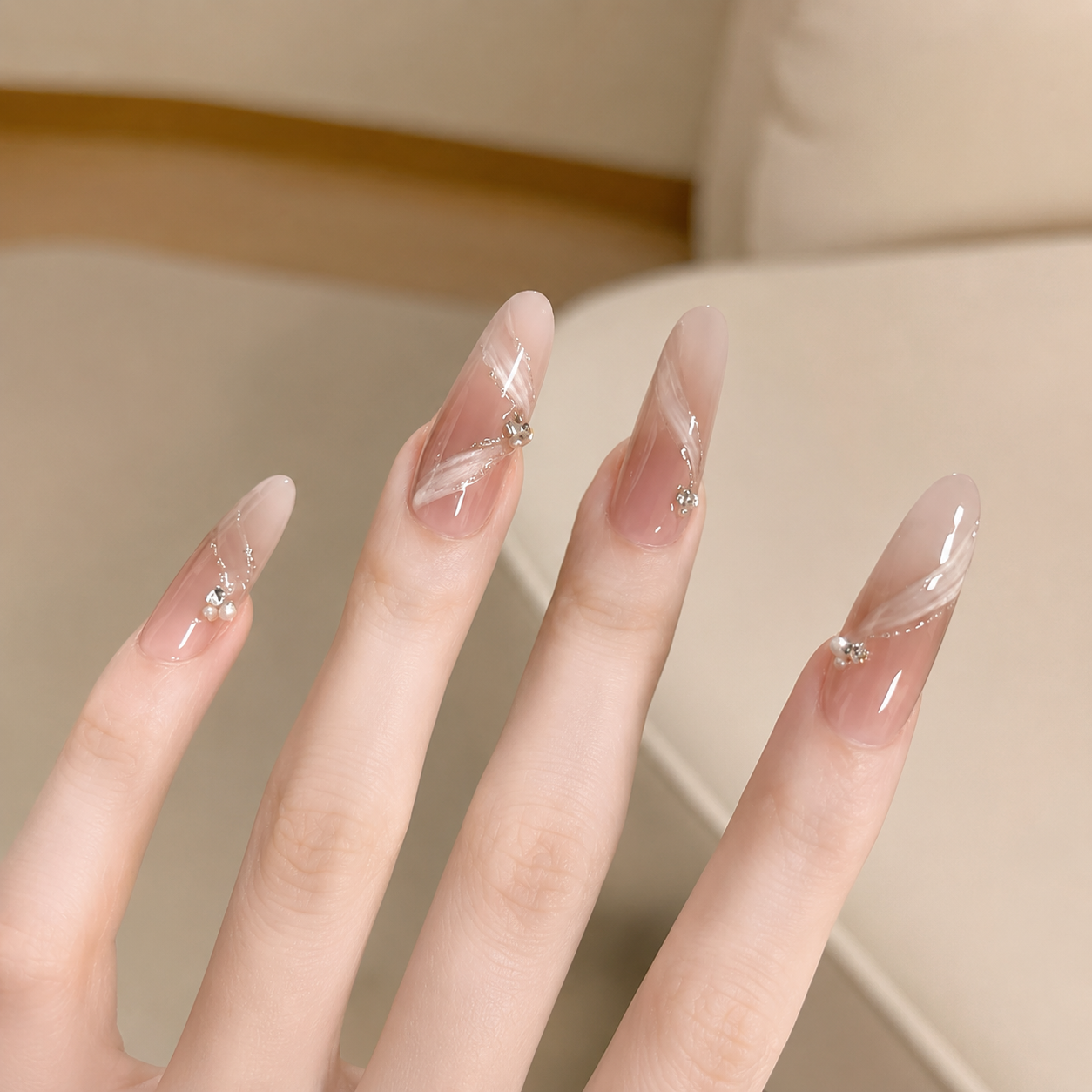 A close-up of a hand with long, almond-shaped nails featuring a sheer nude-pink gradient, white feather-like brushstroke details, and small clear rhinestone accents, set against a soft beige indoor background.