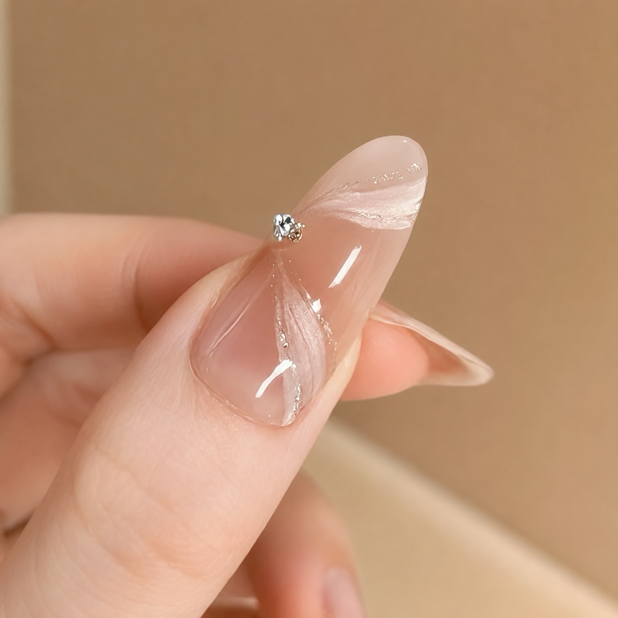 A close-up of a hand showing a long, almond-shaped press-on nail with a glossy nude-pink base, white feather-like streaks, and a small clear rhinestone at the tip, set against a soft beige background.