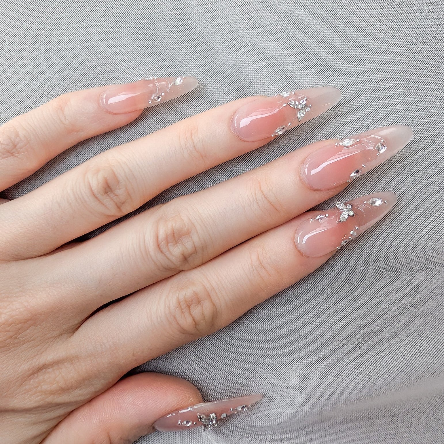 A close-up of a hand with long, almond-shaped nails decorated with a sheer nude-pink base, glossy finish, and small silver rhinestone accents, resting on a light gray textured fabric.