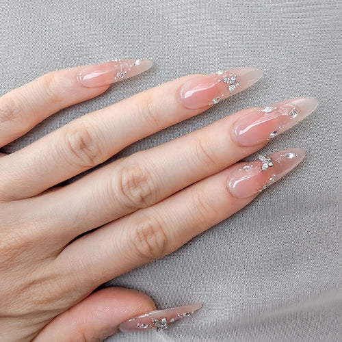 A close-up of a hand with long, almond-shaped nails decorated with a sheer nude-pink base, glossy finish, and small silver rhinestone accents, resting on a light gray textured fabric.