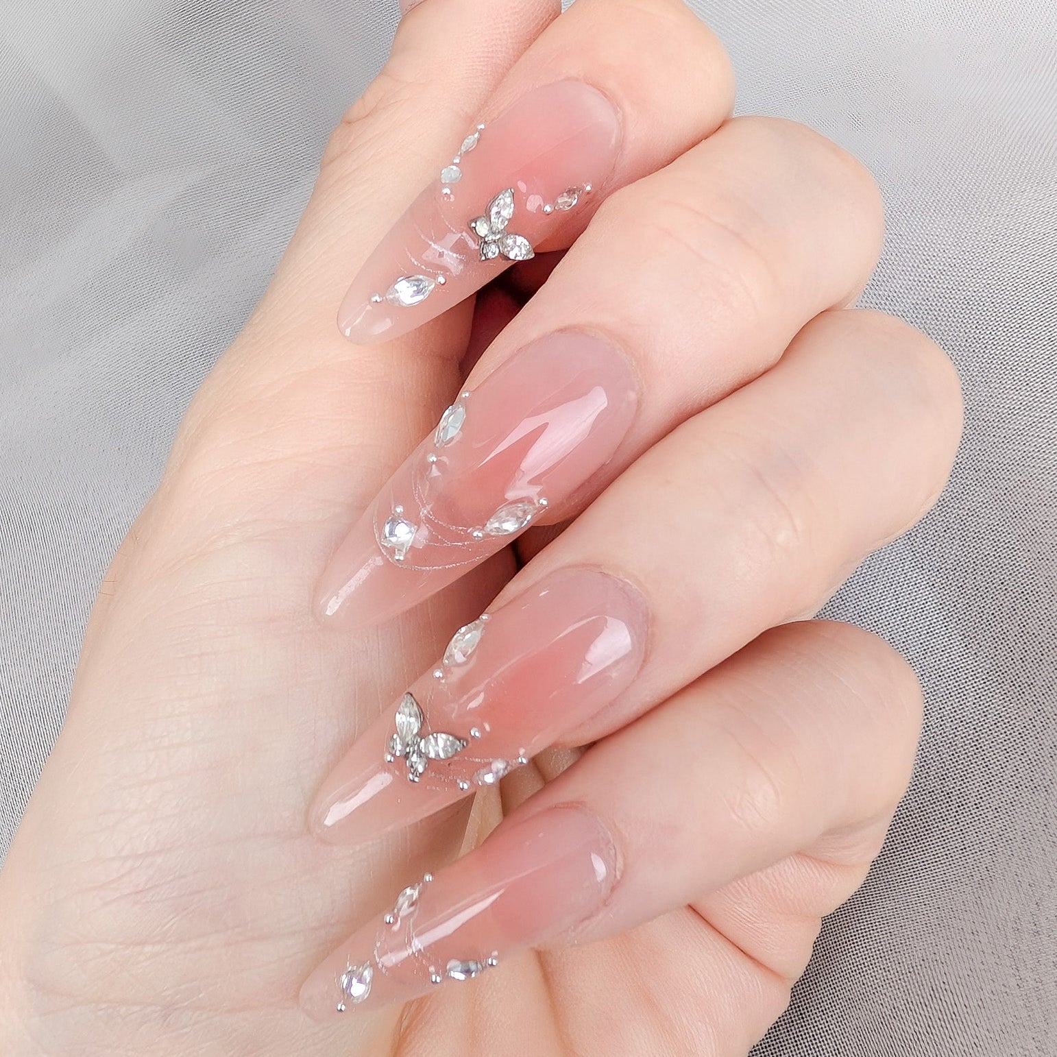 A close-up of a hand with long, almond-shaped nails decorated with a glossy sheer pink base and delicate silver rhinestone clusters, set against a soft gray fabric background.